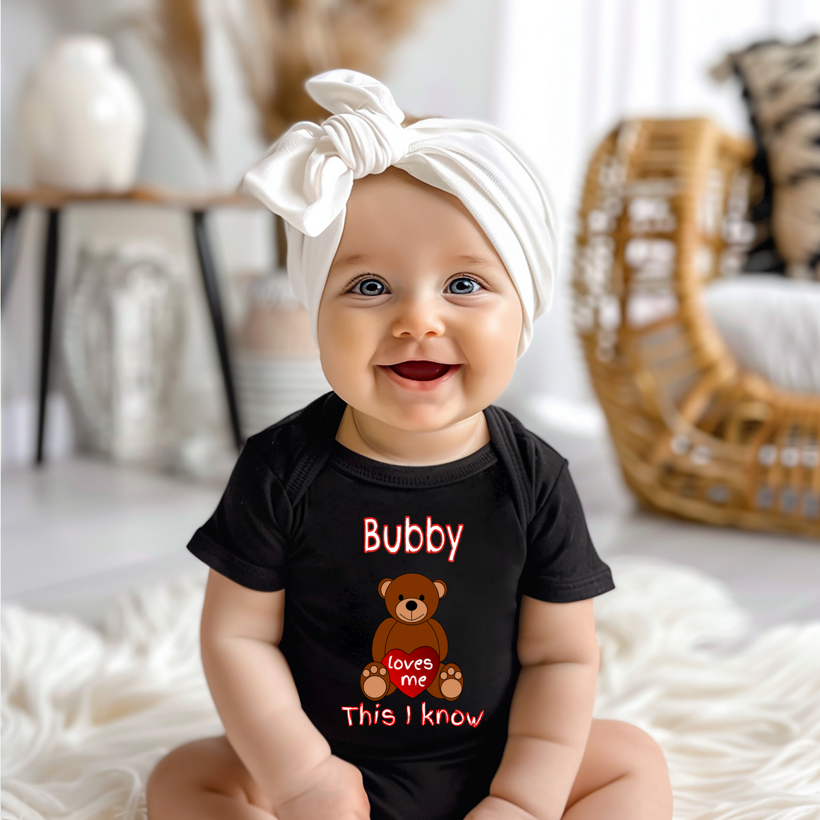 A smiling baby girl wearing a black t-shirt with a teddy bear graphic and the text "Bubby loves me This I know" is sitting on a white blanket.