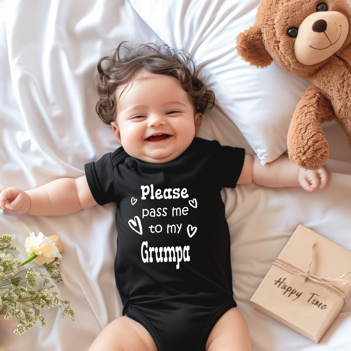 A smiling baby wearing a black onesie with the text "Please pass me to my Grumpa" lies on a bed next to a teddy bear and a gift box.