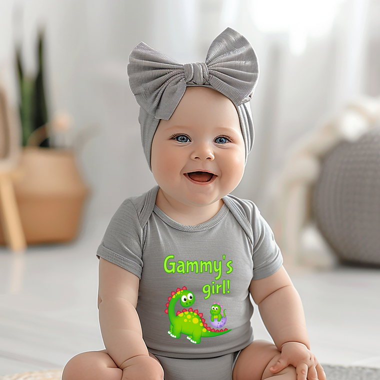 A smiling baby girl wearing a gray headband and a gray onesie with a dinosaur graphic, sitting on a white surface.