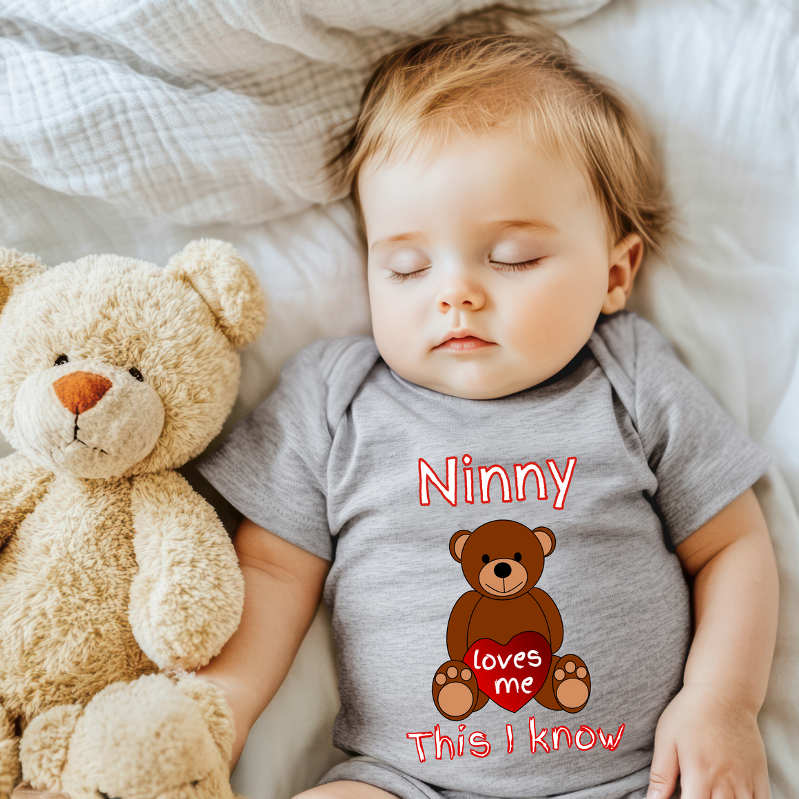 A sleeping baby wearing a gray onesie with a teddy bear and the text "Ninny loves me" printed on it.