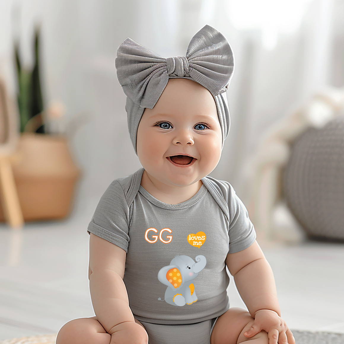 A smiling baby girl wearing a gray headband and a gray onesie with an elephant design, sitting on a white floor.