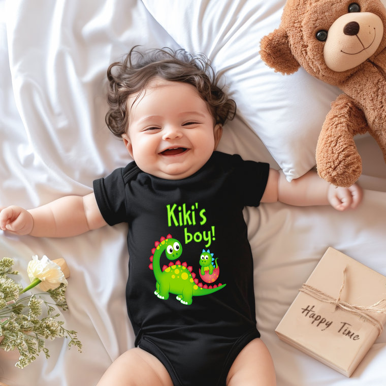 A smiling baby wearing a black onesie with a dinosaur design lies on a bed next to a stuffed teddy bear.