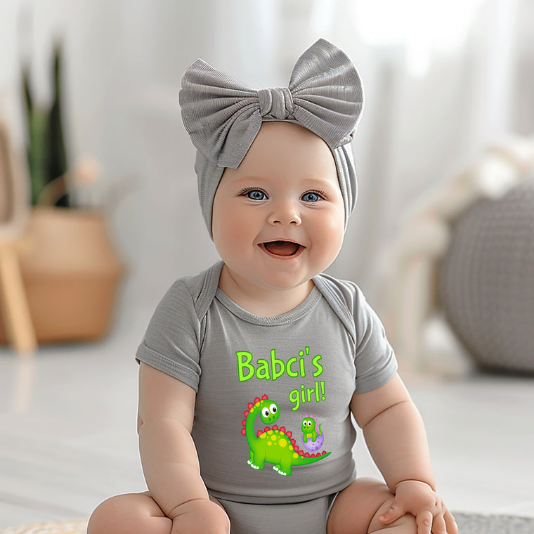 A smiling baby girl wearing a gray headband and a gray onesie with a green dinosaur on it, sitting on a white surface.