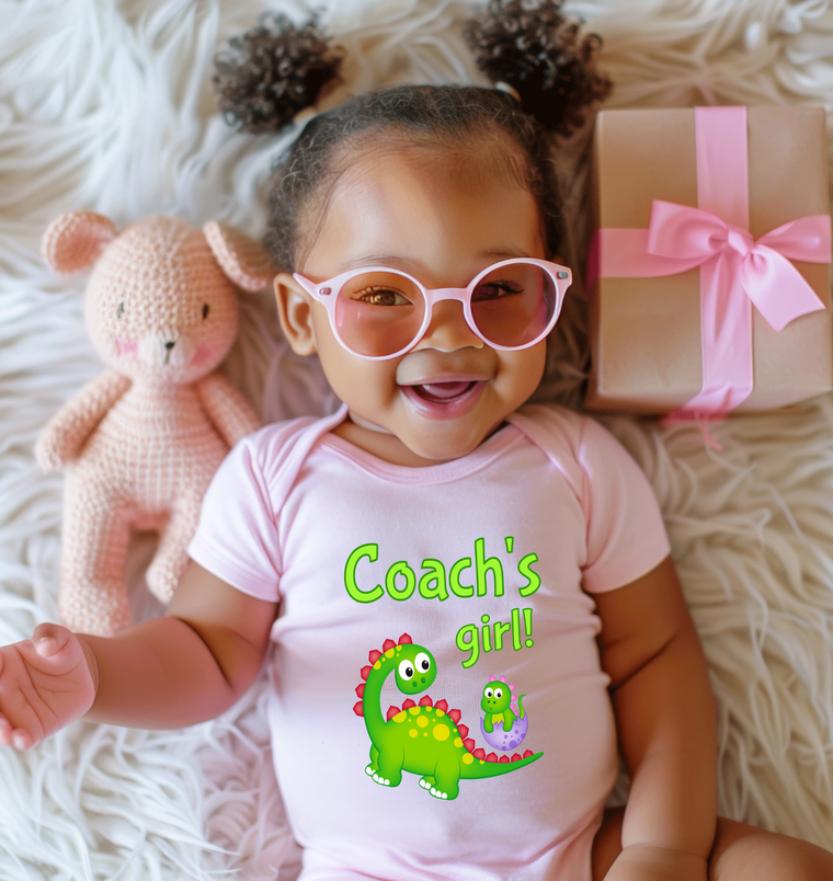 A smiling baby girl wearing a pink outfit with a dinosaur graphic and pink glasses, lying on a white surface next to a pink stuffed animal and a gift box.
