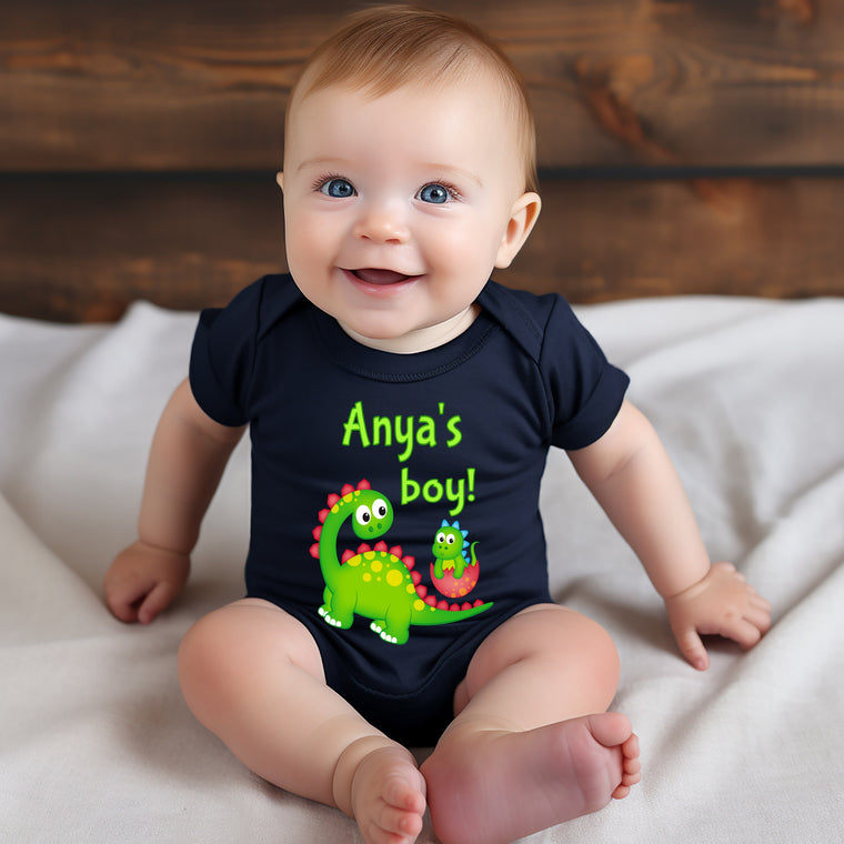 A smiling baby wearing a black shirt with a dinosaur design and the text "Anya's boy!"