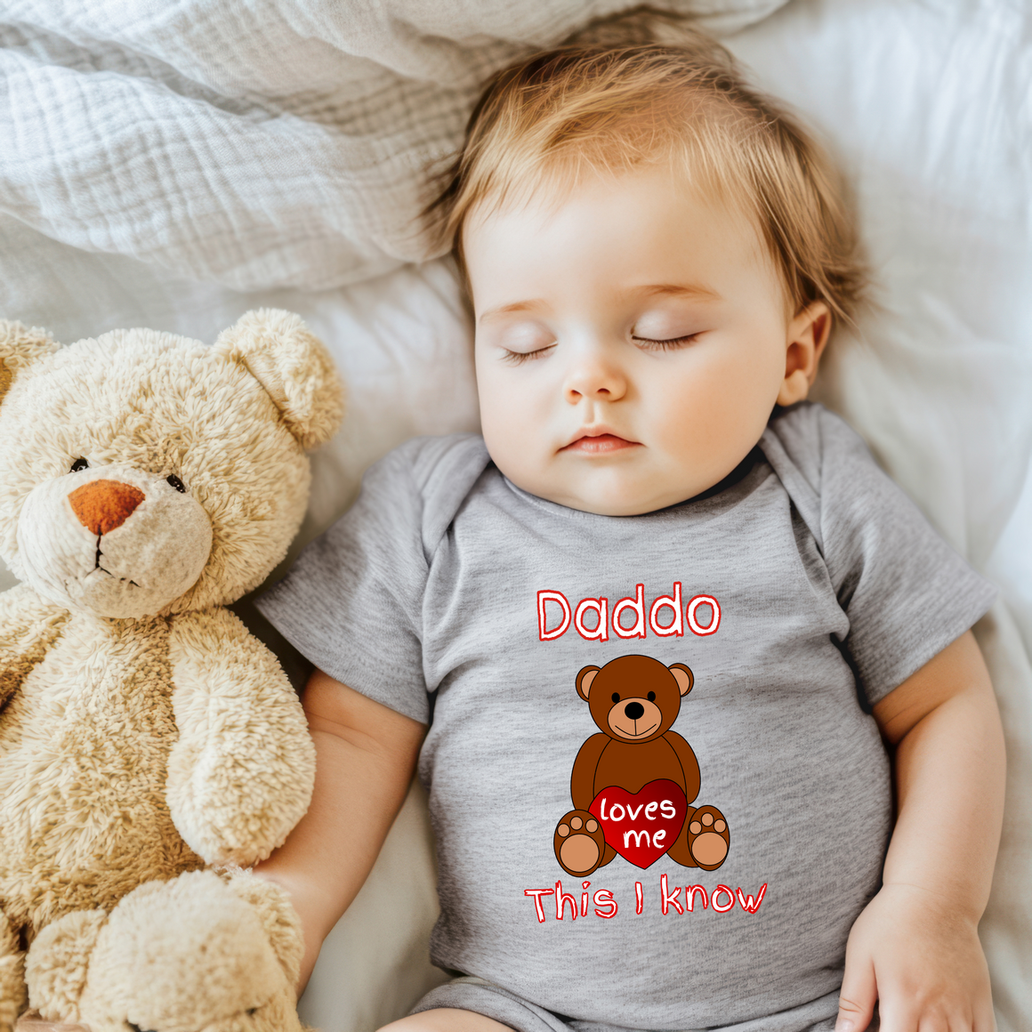 A sleeping baby wearing a grey onesie with a teddy bear and the text "Daddo loves me This I know" printed on it.
