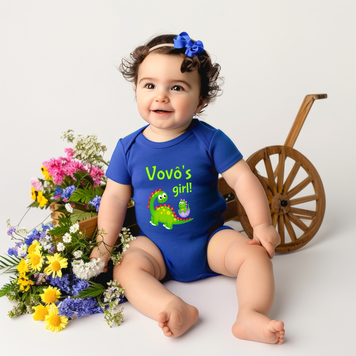 A smiling baby girl wearing a blue onesie with a dinosaur and the text "Vovô's girl!" is sitting on the floor surrounded by a bouquet of colorful flowers and a wooden toy wagon.
