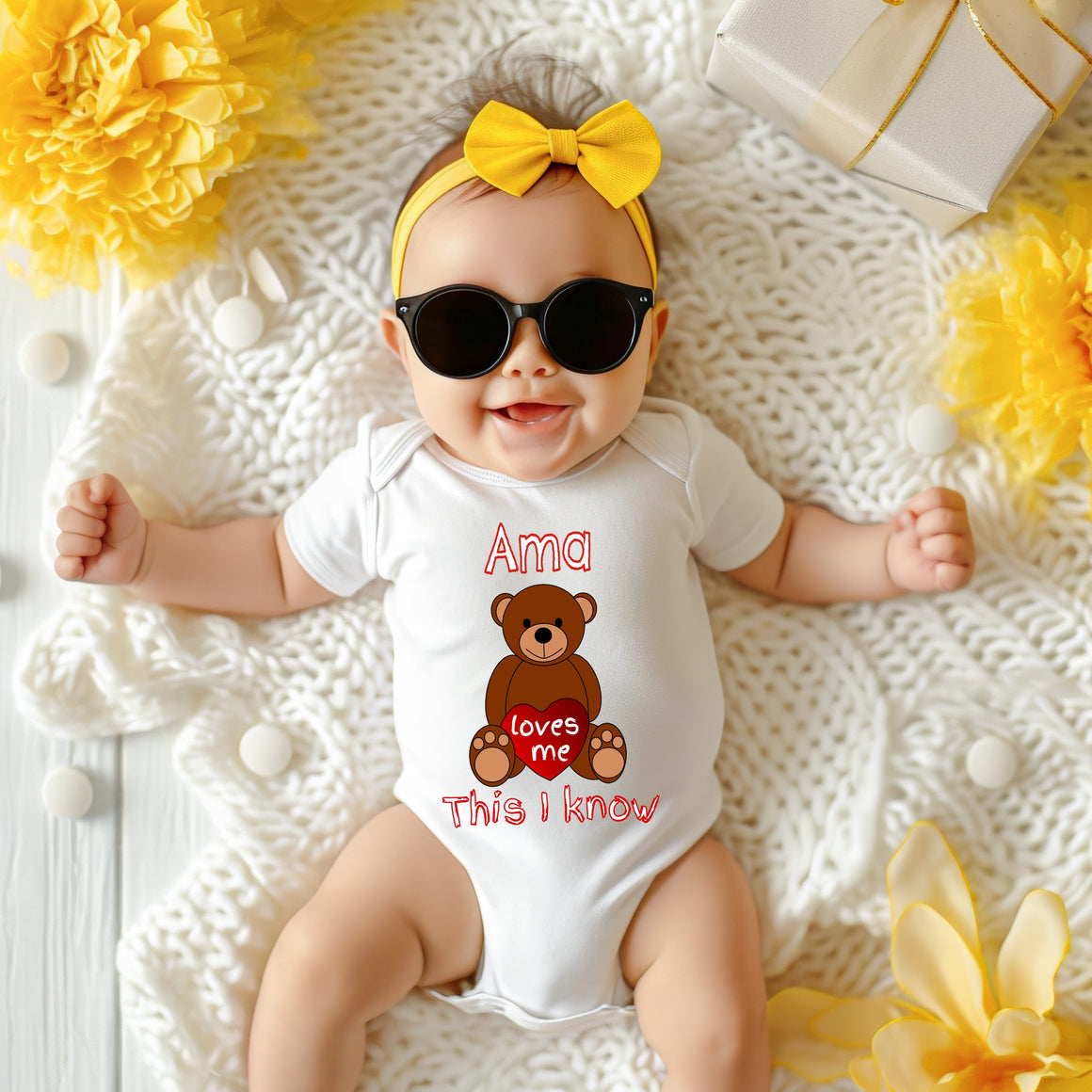 A baby wearing a white onesie with a teddy bear graphic and the text "Ama loves me This I know" is lying on a white blanket surrounded by yellow flowers.