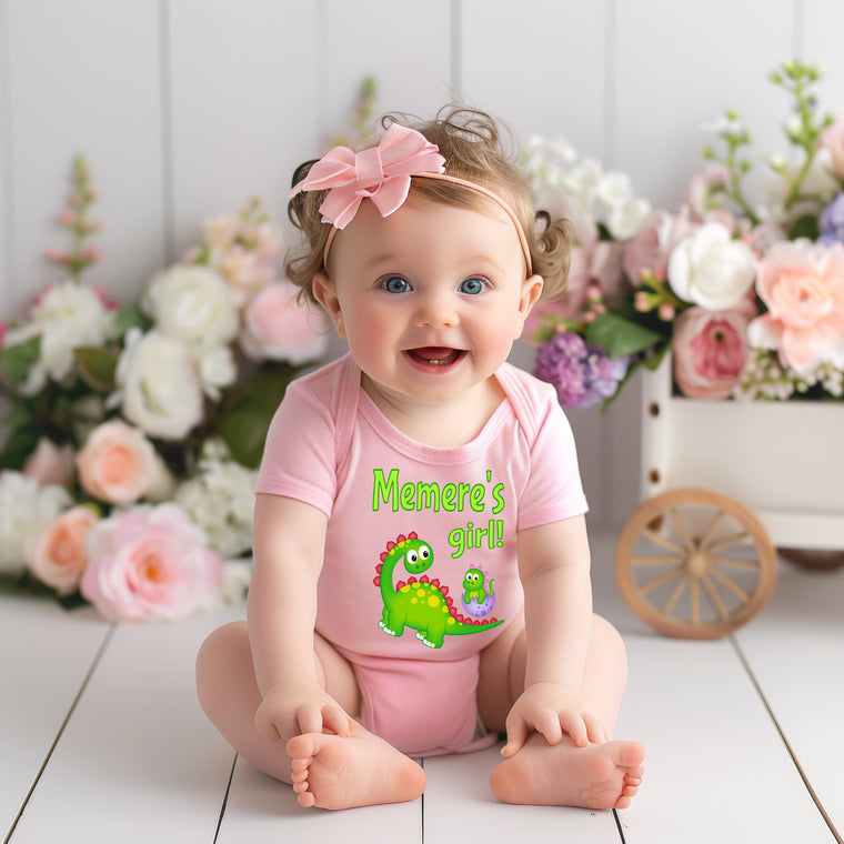 A smiling baby girl wearing a pink outfit with a dinosaur design sits on a wooden floor, surrounded by a bouquet of flowers.