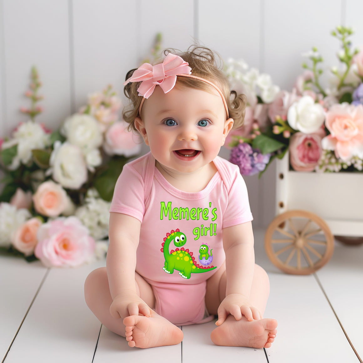 A smiling baby girl wearing a pink outfit with a dinosaur design sits on a wooden floor, surrounded by a bouquet of flowers.