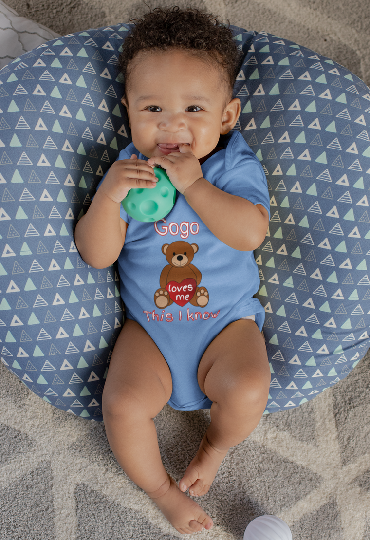A smiling baby wearing a blue onesie with a teddy bear design, lying on a blue and gray patterned pillow.