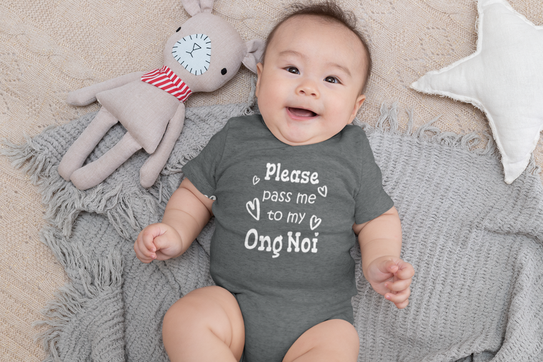 A smiling baby wearing a gray onesie with the text "Please pass me to my Ong Noi" lies on a gray blanket next to a stuffed animal.