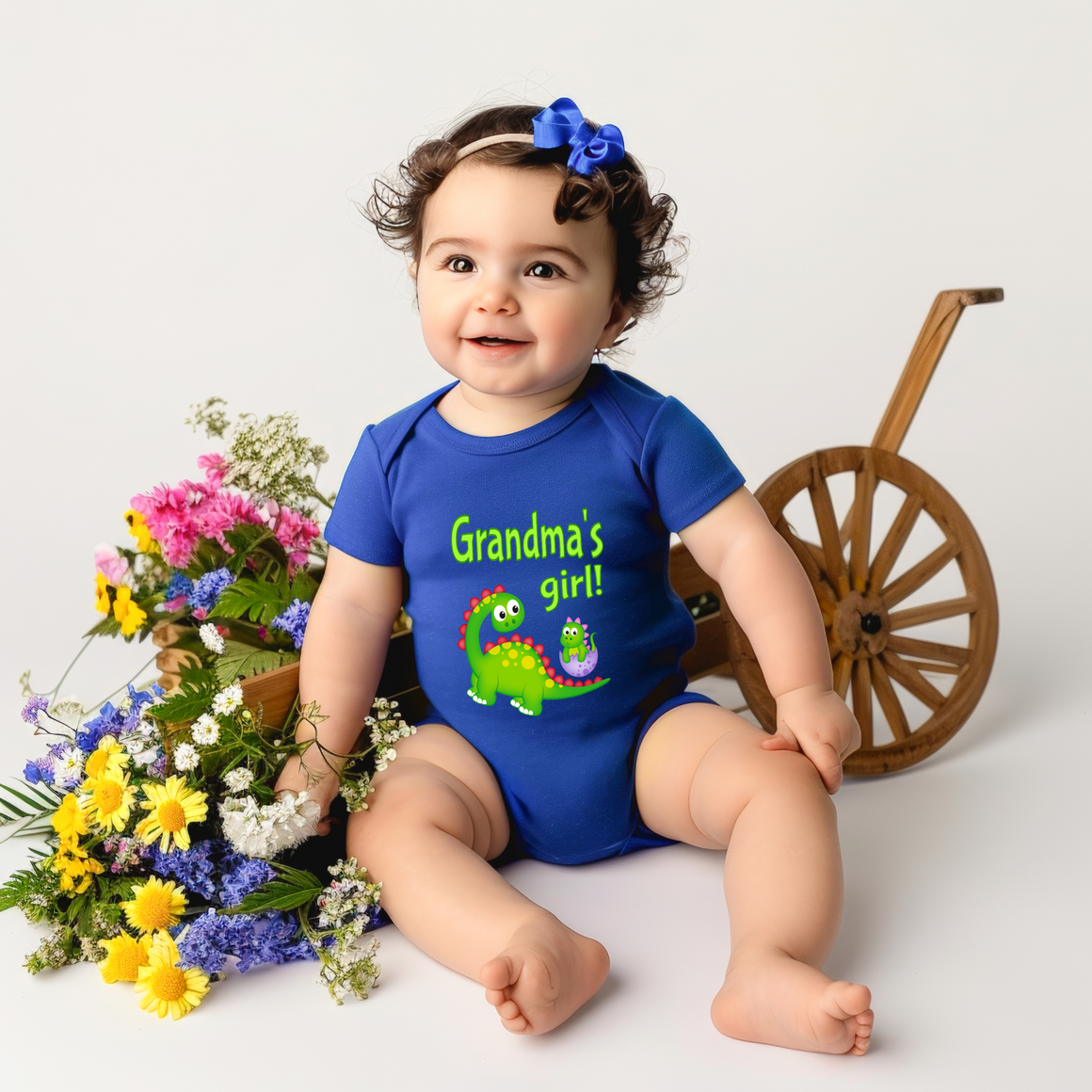 A smiling baby girl wearing a blue onesie with a dinosaur and the text "Grandma's girl!" is sitting next to a wooden wagon wheel and a basket filled with colorful flowers.