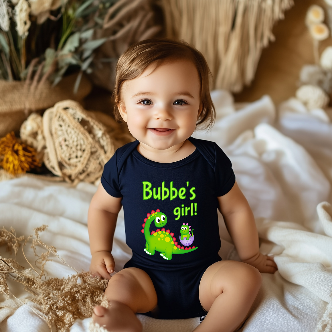 A smiling baby boy wearing a black t-shirt with a dinosaur graphic and the text "Bubbe's girl!"