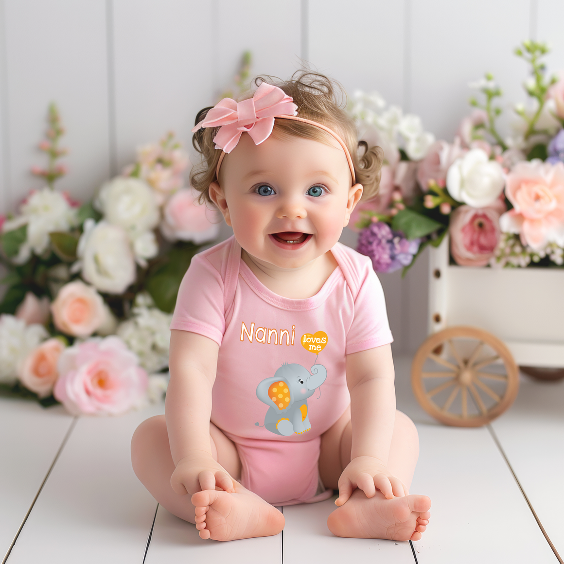 A smiling baby girl wearing a pink outfit with a cartoon elephant on it, sitting on a white wooden floor in front of a bouquet of flowers.