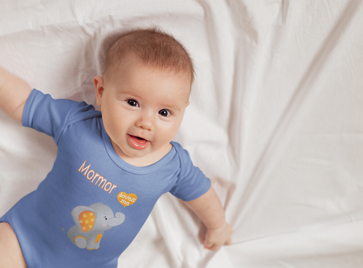 A smiling baby wearing a blue onesie with an elephant design lies on a white sheet.