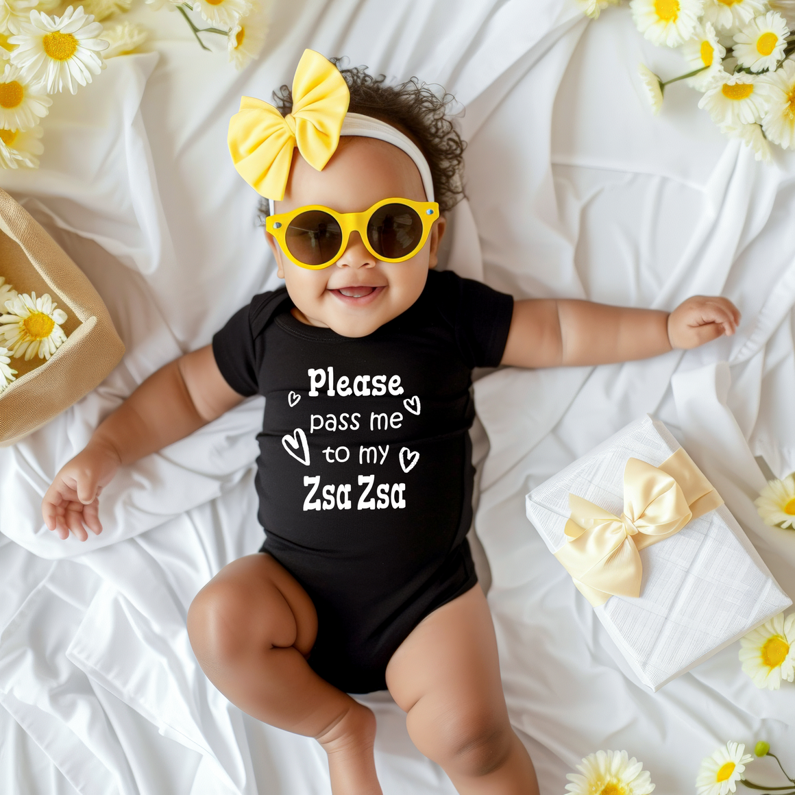 A smiling baby girl wearing a black outfit with a yellow bow and sunglasses lies on a white sheet, surrounded by yellow flowers.