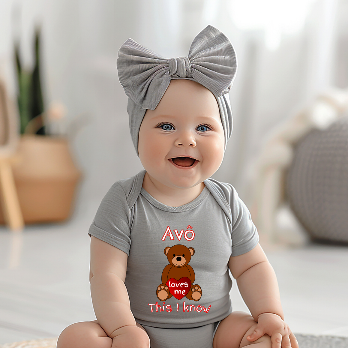 A smiling baby girl wearing a gray headband and a gray onesie with a teddy bear graphic and text.