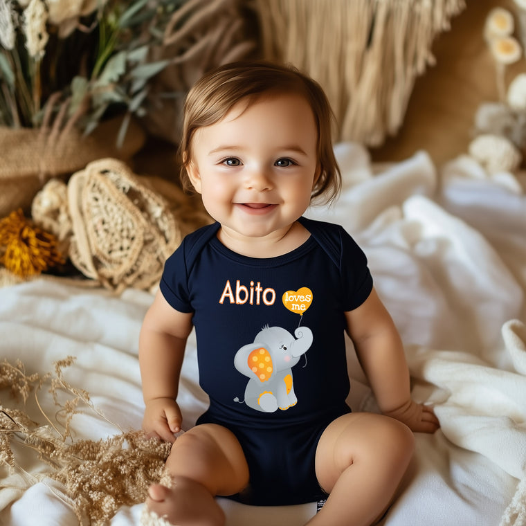 A smiling baby boy wearing a navy blue onesie with an elephant design sits on a bed surrounded by various objects.