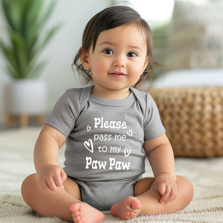 A smiling baby girl wearing a gray onesie with the text "Please pass me to my paw" sits on a beige rug.