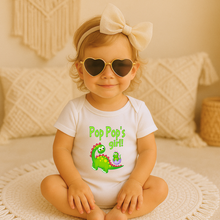 A young girl wearing a white t-shirt with a cartoon dinosaur and the text "Pop Pop's girl!" sits on a bed, smiling and wearing heart-shaped sunglasses.