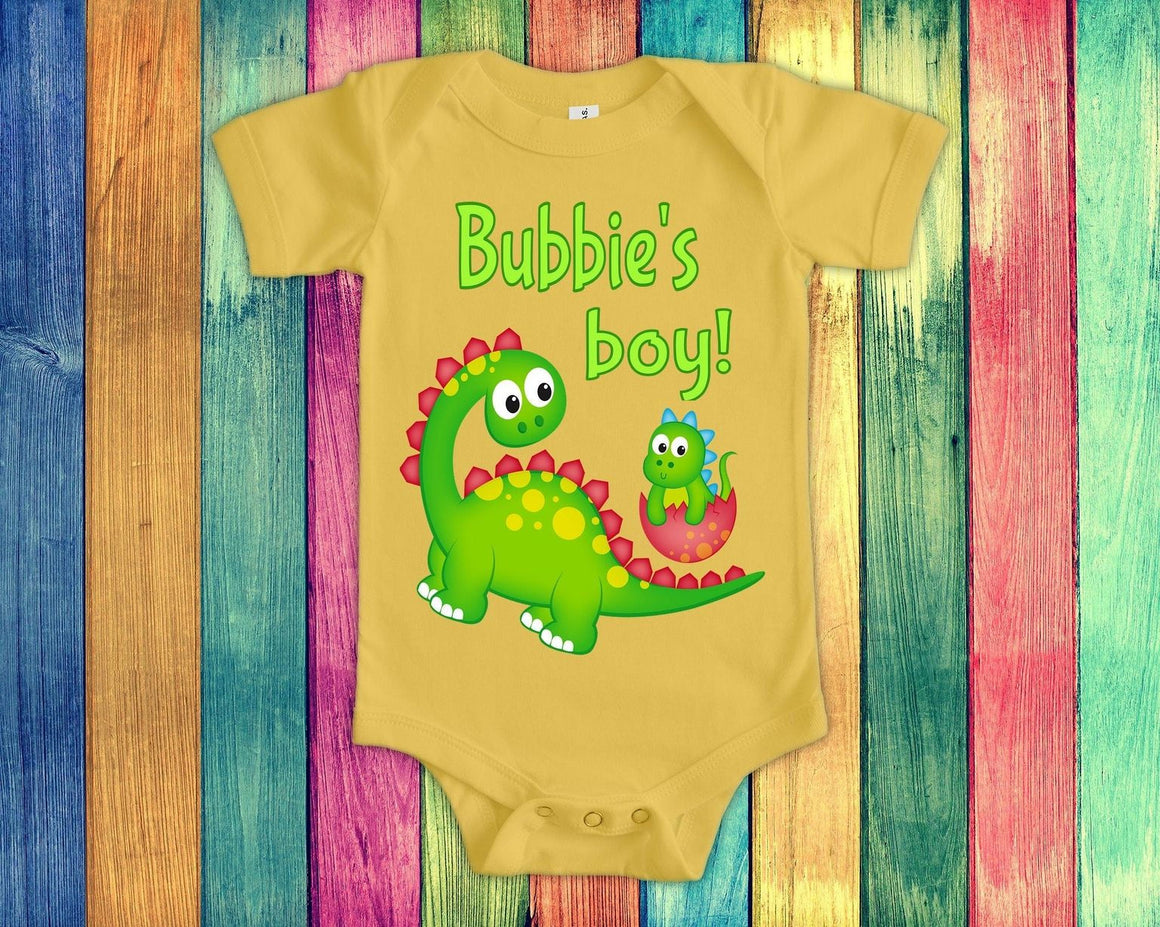 A yellow baby onesie with a cartoon dinosaur and the text "Bubbie's boy!" printed on it, placed on a colorful wooden background.
