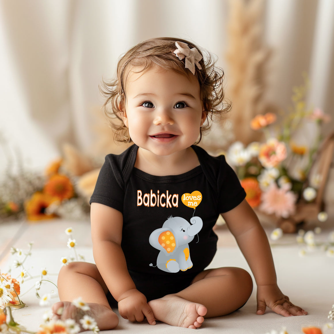 A smiling baby girl wearing a black t-shirt with an elephant design sits on the floor surrounded by flowers.