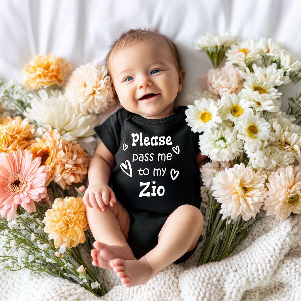 A smiling baby wearing a black onesie with the text "Please pass me to my Zio" is surrounded by a bouquet of colorful flowers.