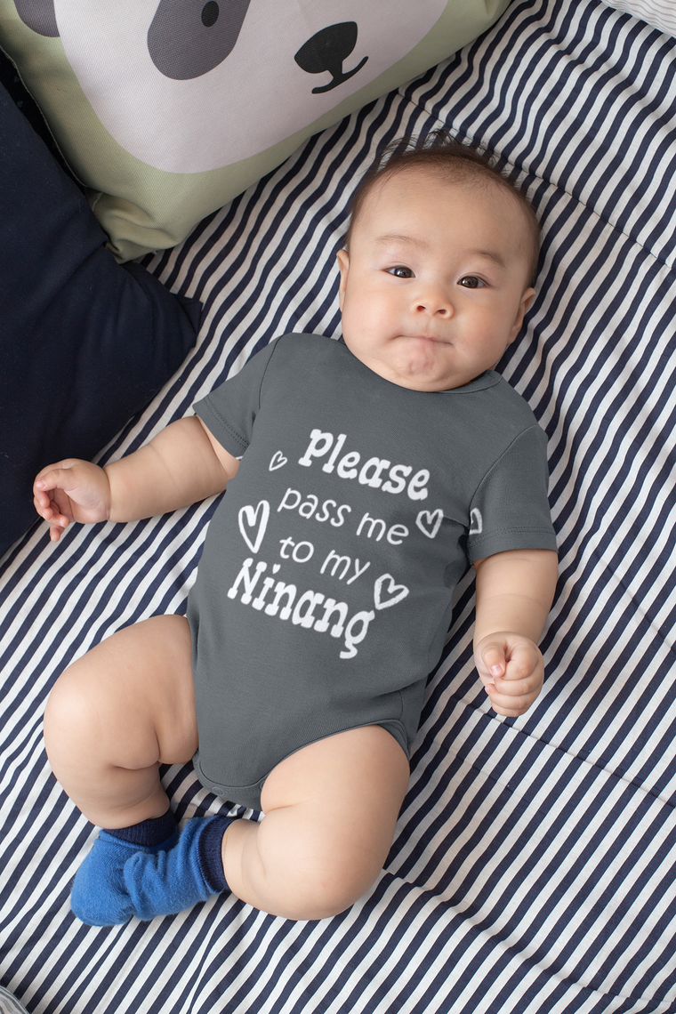 A baby wearing a grey onesie with the text "Please pass me to my Ninang" lying on a striped bed.