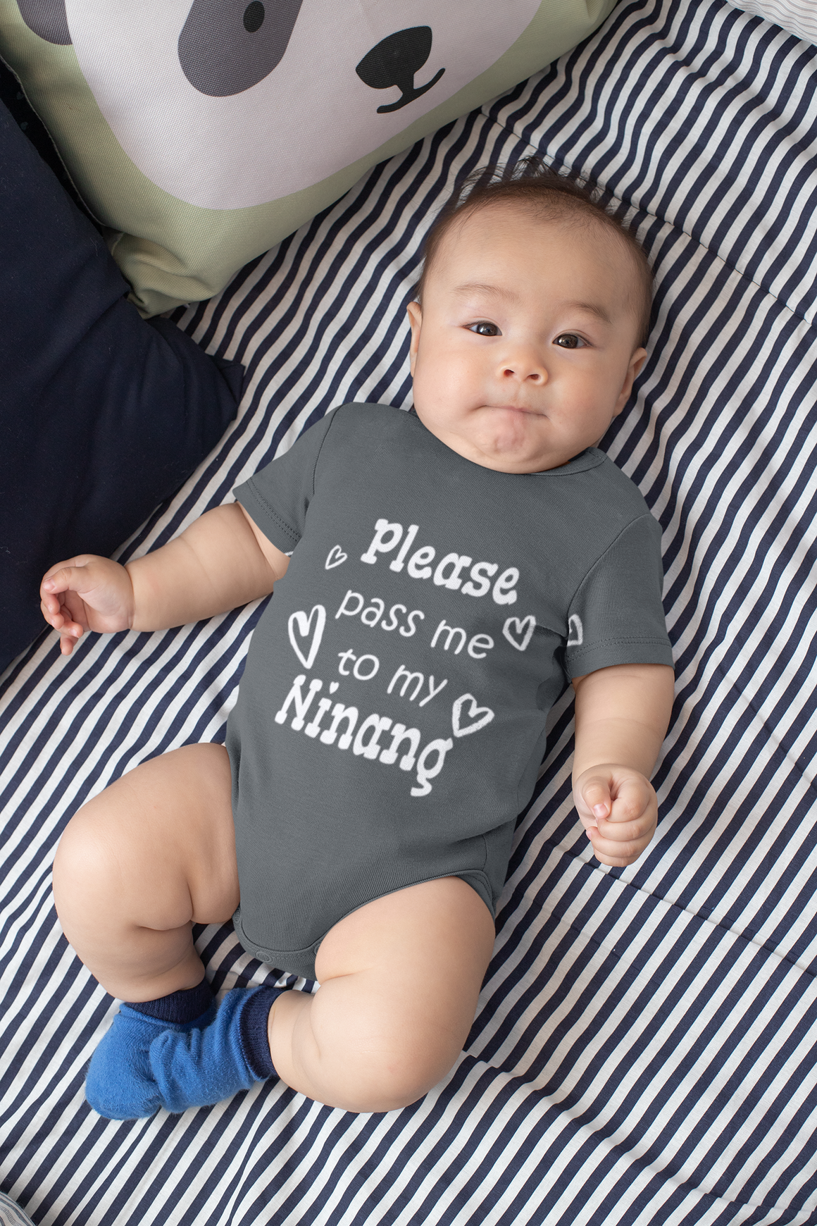 A baby wearing a grey onesie with the text "Please pass me to my Ninang" lying on a striped bed.