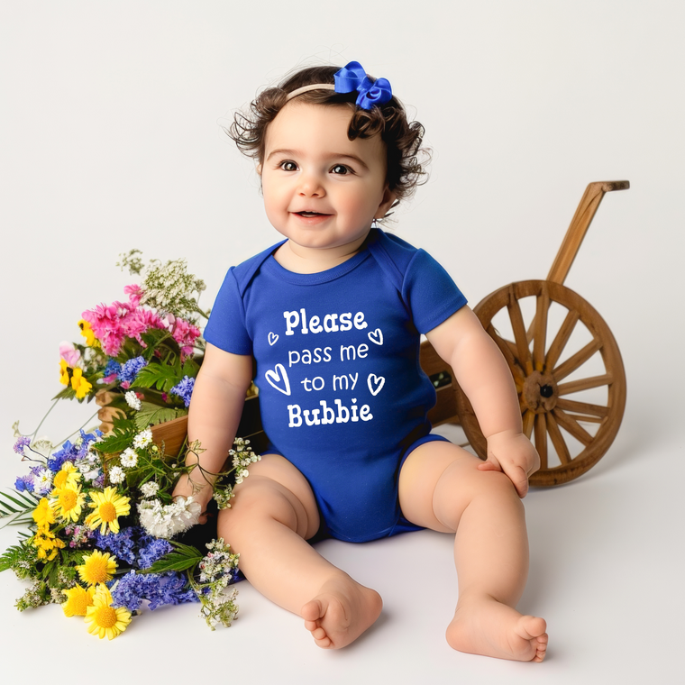 A smiling baby girl wearing a blue onesie with the text "Please pass me to my bubble" sits next to a wooden wagon filled with colorful flowers.