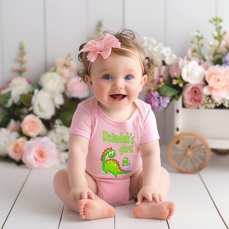 A smiling baby girl wearing a pink outfit with a dinosaur design sits on a wooden floor, surrounded by a bouquet of flowers.