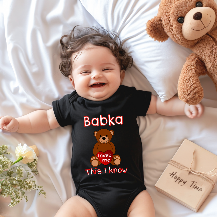 A smiling baby wearing a black onesie with a teddy bear and the text "Babka loves me This I know" lies on a bed next to a stuffed teddy bear and a book.