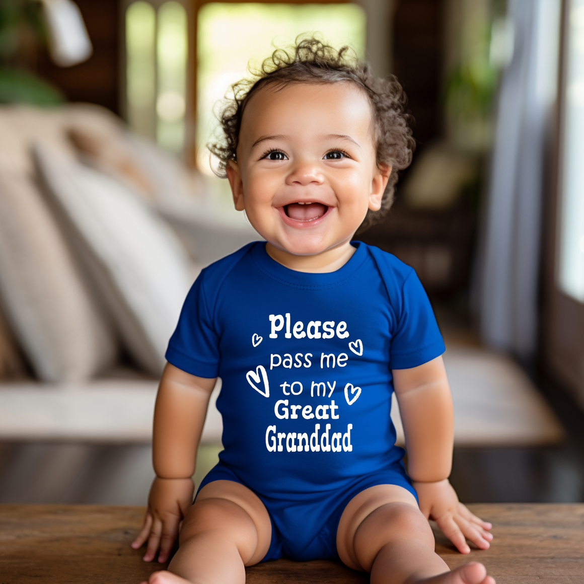 A smiling baby boy wearing a blue onesie with the text "Please pass me to my Great Granddad" is sitting on a wooden surface.