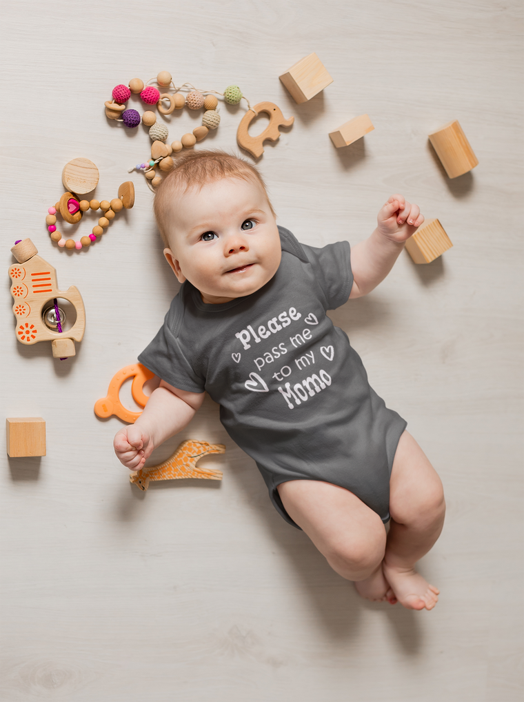 A baby lies on a white floor surrounded by various toys, including wooden blocks, a toy train, and a giraffe-shaped teething ring. The baby is wearing a gray onesie with the text "Please pass me to my Momo" printed on it.