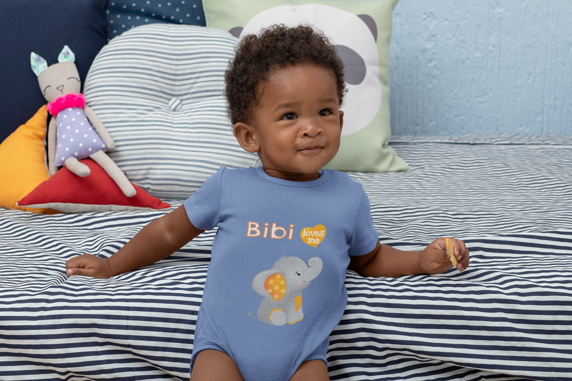 A young child wearing a blue onesie with an elephant design sits on a bed surrounded by colorful pillows.