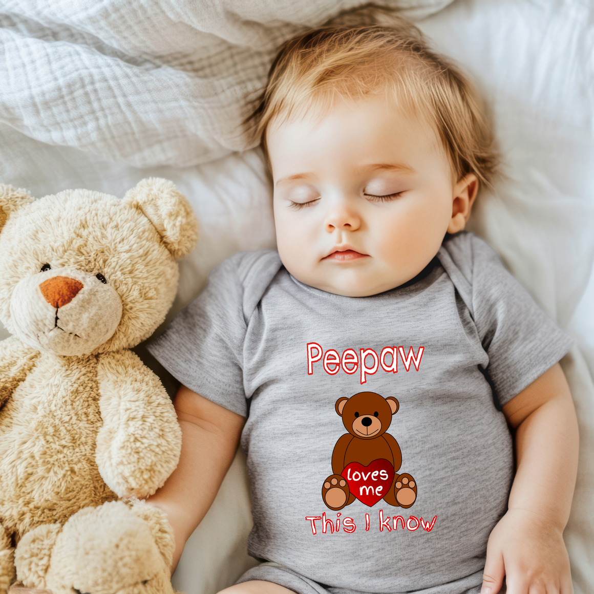 A sleeping baby wearing a grey shirt with a teddy bear and the text "Peepaw loves me This I know" is lying on a bed.