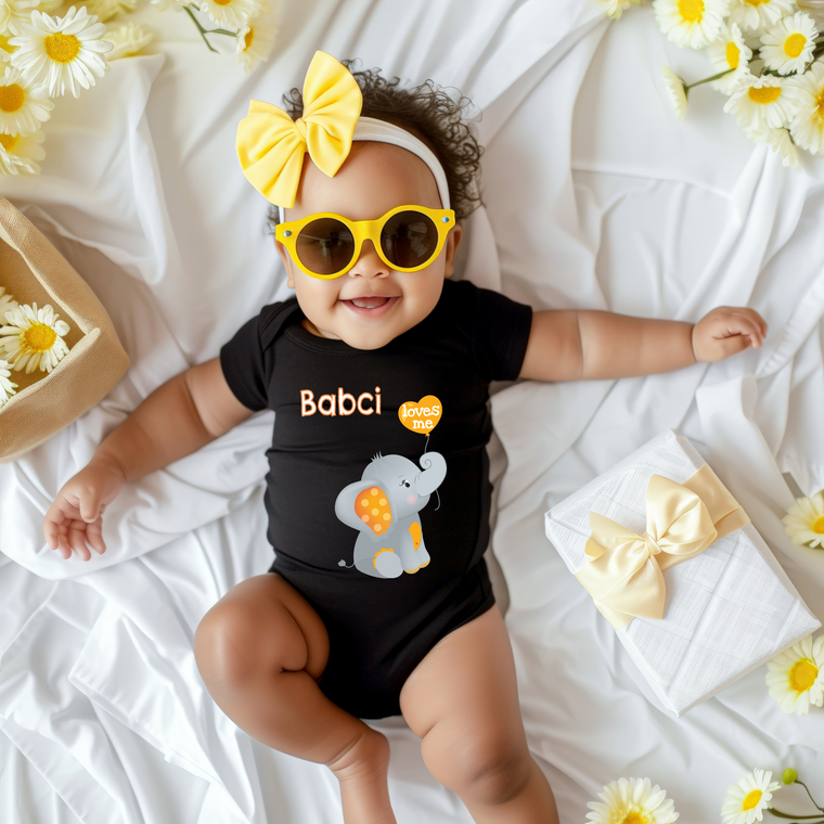 A smiling baby girl wearing a black outfit with a yellow bow and yellow sunglasses lies on a white sheet, surrounded by yellow flowers.
