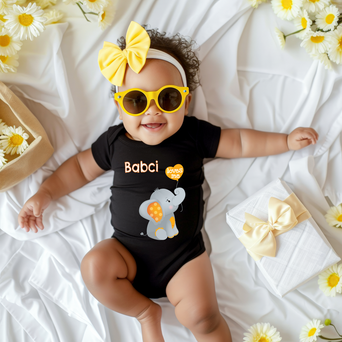 A smiling baby girl wearing a black outfit with a yellow bow and yellow sunglasses lies on a white sheet, surrounded by yellow flowers.