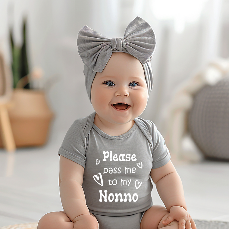 A smiling baby girl wearing a gray headband and a gray onesie with the text "Please pass me to my Nonno" is sitting on the floor.