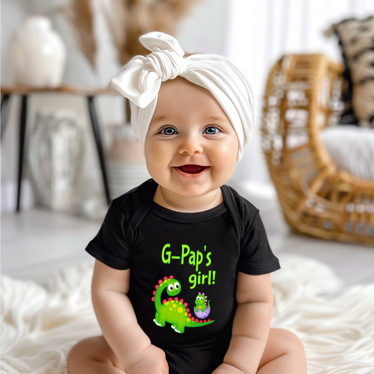 A smiling baby girl wearing a black t-shirt with a dinosaur graphic and text, sitting on a white surface.
