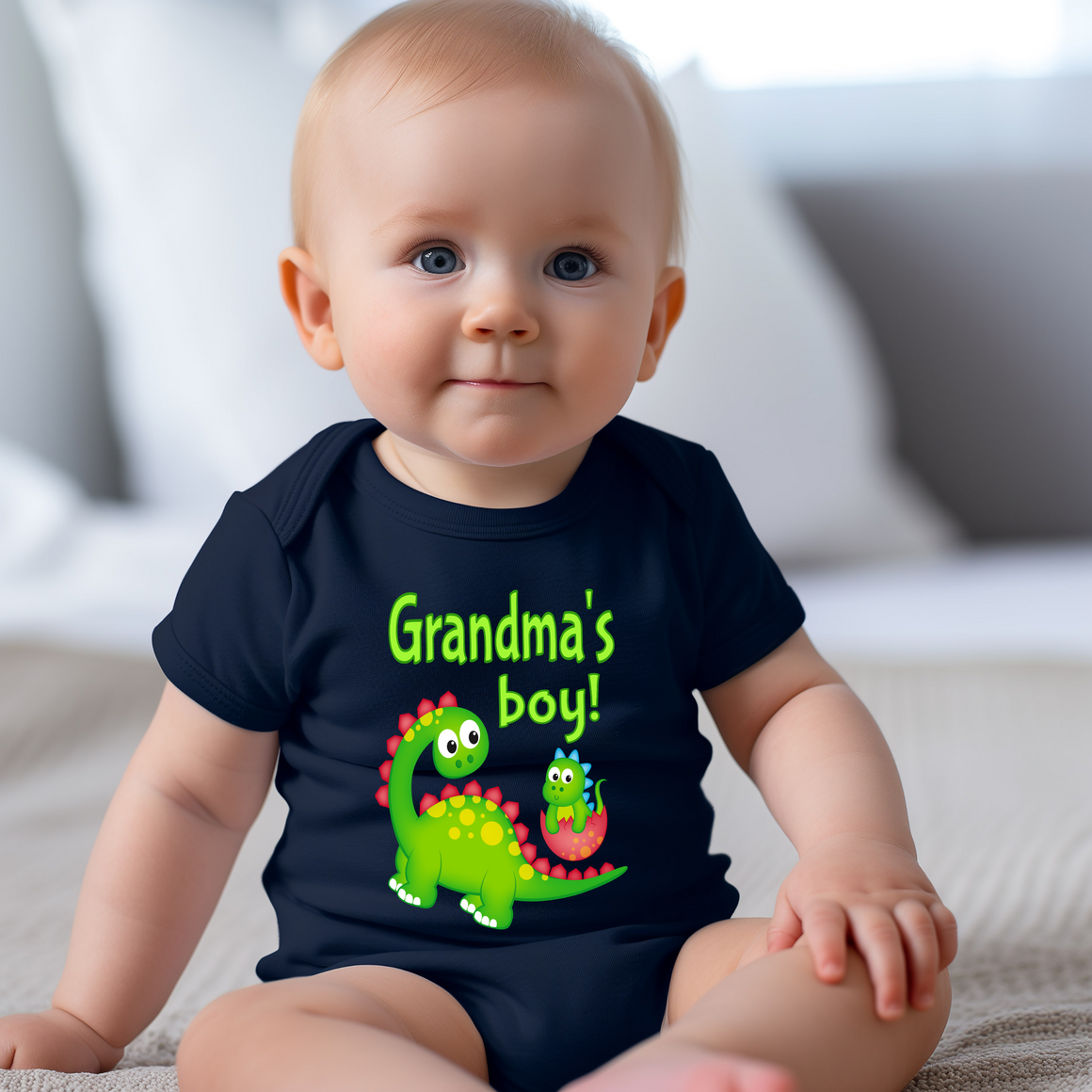 A smiling baby wearing a black t-shirt with a dinosaur graphic and text that says "Grandma's boy!".