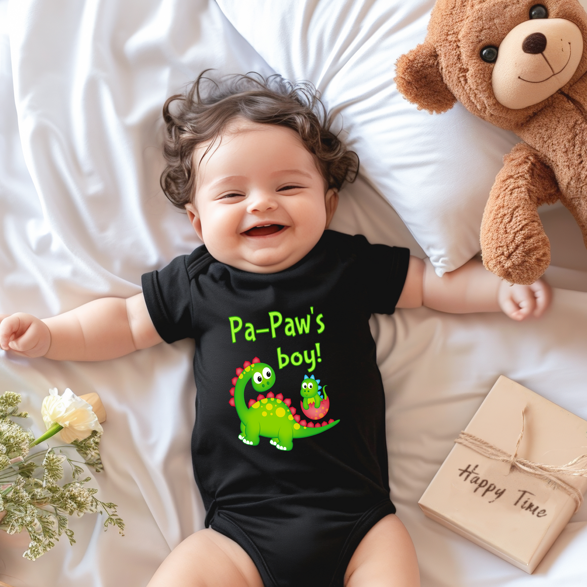 A smiling baby wearing a black t-shirt with a dinosaur graphic lies on a bed next to a stuffed teddy bear.