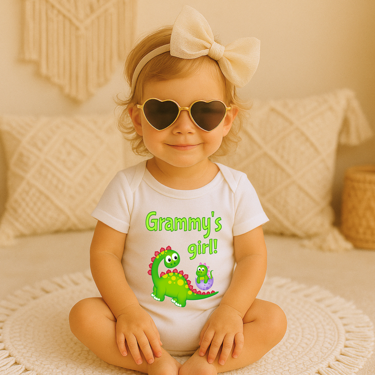 A young girl wearing a white t-shirt with a dinosaur graphic and the text "Granny's girl!" sits on a white rug, smiling and wearing heart-shaped sunglasses.