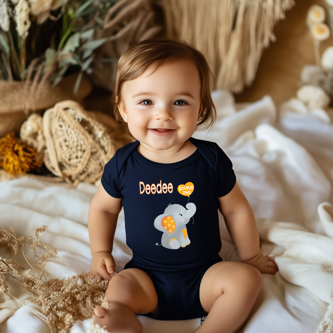 A smiling baby boy wearing a navy blue onesie with an elephant design sits on a white blanket.