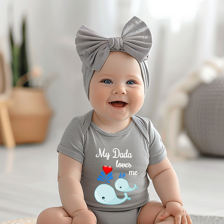 A smiling baby girl wearing a gray headband and a gray onesie with a heart and whale design, sitting on a white surface.