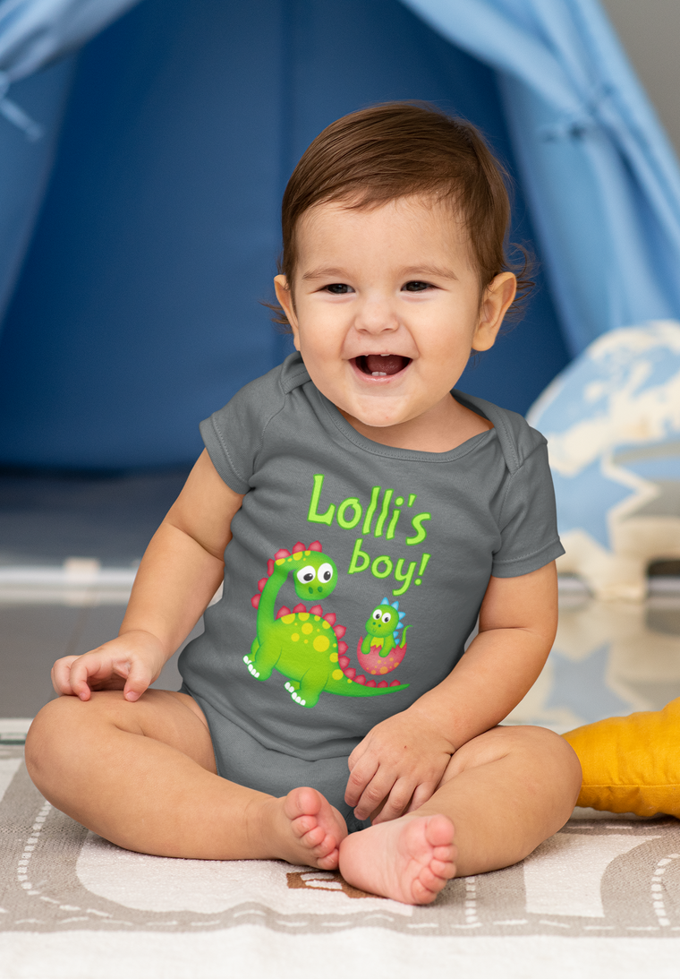 A smiling baby boy wearing a gray onesie with a green dinosaur and text on it, sitting on a gray and white rug.