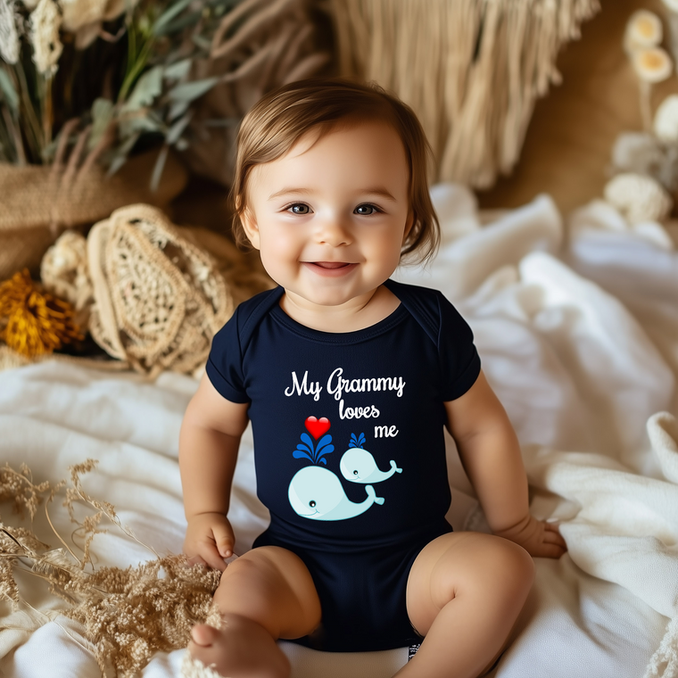 A smiling baby wearing a navy blue onesie with a whale design and the text "My Grammy loves me" is sitting on a bed.