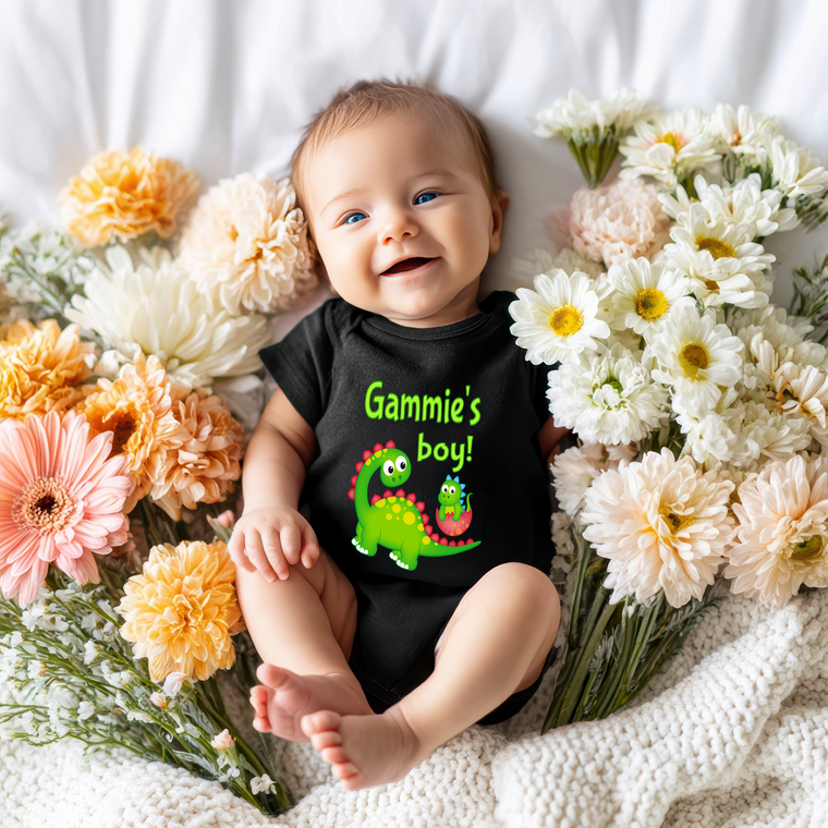 A smiling baby wearing a black shirt with a dinosaur graphic lies on a bed surrounded by colorful flowers.