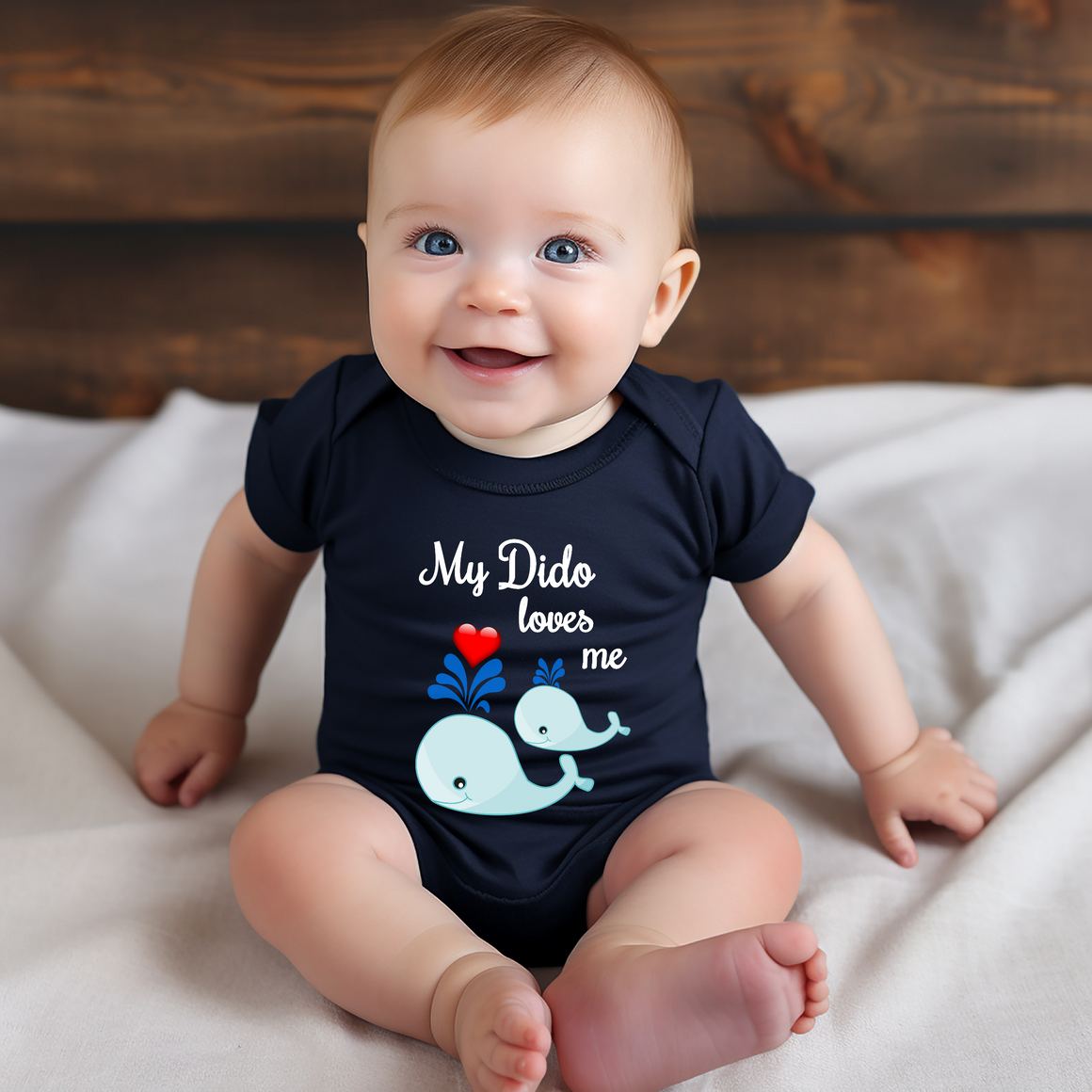 A smiling baby wearing a navy blue onesie with a cartoon whale design and the text "My Dido loves me" printed on it.